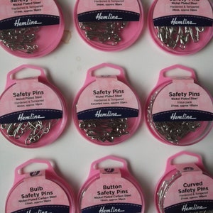 May include: Nine pink packages of Hemline safety pins, each with a clear window showing the nickel-plated steel pins. The labels detail pin sizes in mm and approximate quantities. Styles include bulb, button, and curved safety pins.