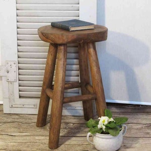 Könnte beinhalten: Ein runder, dunkelbrauner Holzhocker mit vier stabilen Beinen und einer kreisförmigen Sitzfläche. Ein kleines, dunkel gefärbtes Buch liegt flach in der Mitte der Sitzfläche. Der Hocker hat eine natürliche Holzmaserung und eine einfache Konstruktion.
