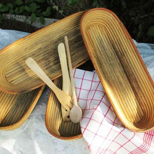 Op de afbeelding: Drie geweven, ovale broodmanden gemaakt van natuurlijk hout in een lichtbruine kleur. De manden zijn op elkaar gestapeld en staan op een wit tafelkleed. Er liggen twee houten lepels naast de manden.
