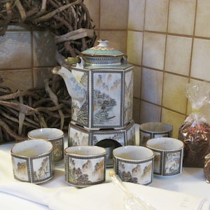 Puede incluir: Un juego de té de estilo asiático vintage con una tetera, seis tazas y un soporte para calentar. El juego presenta ilustraciones de paisajes en tonos apagados. También hay dos bolsas de celofán con té.