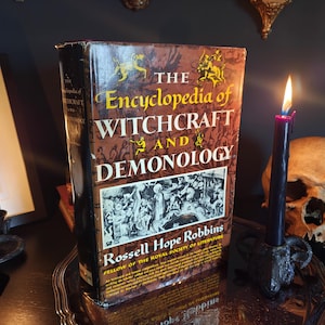 May include: A hardcover book titled "The Encyclopedia of Witchcraft and Demonology" stands upright on a silver tray. The book has a brown cover with gold lettering and illustrations. A lit black candle and a skull are next to the book.