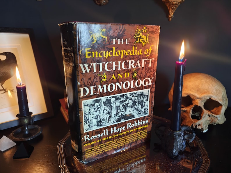 May include: A hardcover book titled "The Encyclopedia of Witchcraft and Demonology" stands upright on a silver tray. The book has a brown cover with gold lettering and illustrations. A lit black candle and a skull are next to the book.