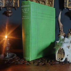 Może przedstawiać: Zielona książka zatytułowana "Witchcraft" z podtytułem "Its power in the World Today" autorstwa Williama Seabrooka. Obok książki znajduje się czaszka jelenia z rogami i zapalona świeca w mosiężnym świeczniku. Scena rozgrywa się na drewnianej powierzchni.