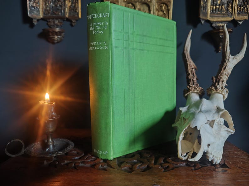 Może przedstawiać: Zielona książka zatytułowana "Witchcraft" z podtytułem "Its power in the World Today" autorstwa Williama Seabrooka. Obok książki znajduje się czaszka jelenia z rogami i zapalona świeca w mosiężnym świeczniku. Scena rozgrywa się na drewnianej powierzchni.