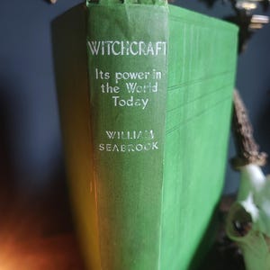 Może przedstawiać: Zabytkowa książka w twardej oprawie w kolorze zielonym zatytułowana "WITCHCRAFT" z podtytułem "Its power in the World Today" autorstwa Williama Seabrooka. Grzbiet książki ma białe litery. Książka jest prezentowana na ciemnej powierzchni.
