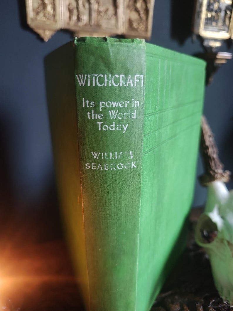 Może przedstawiać: Zabytkowa książka w twardej oprawie w kolorze zielonym zatytułowana "WITCHCRAFT" z podtytułem "Its power in the World Today" autorstwa Williama Seabrooka. Grzbiet książki ma białe litery. Książka jest prezentowana na ciemnej powierzchni.
