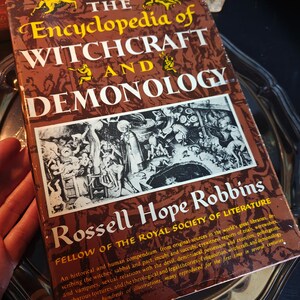 May include: A vintage book titled "The Encyclopedia of Witchcraft and Demonology" by Rossell Hope Robbins. The cover is brown with gold lettering and illustrations. The book is an historical compendium on witchcraft and related topics.