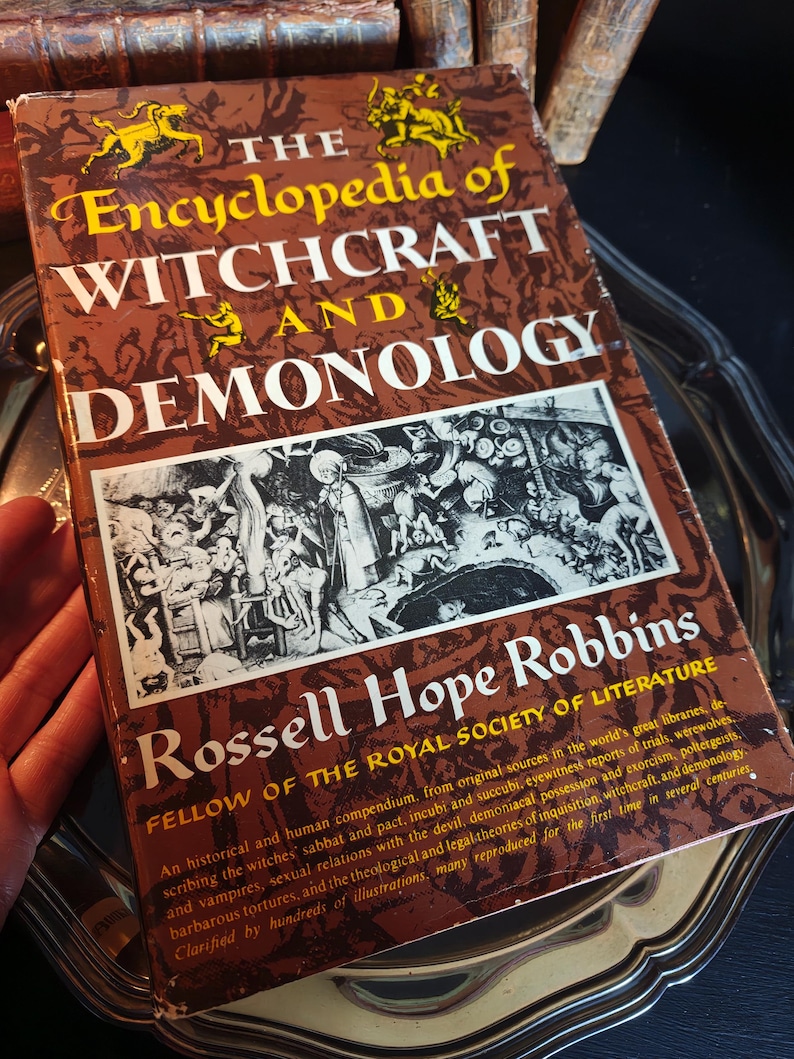 May include: A vintage book titled "The Encyclopedia of Witchcraft and Demonology" by Rossell Hope Robbins. The cover is brown with gold lettering and illustrations. The book is an historical compendium on witchcraft and related topics.