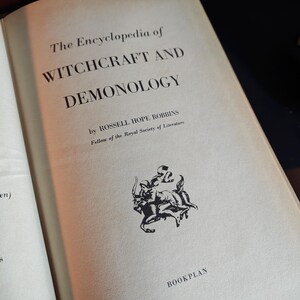 May include: Open book titled "The Encyclopedia of Witchcraft and Demonology" by Rossell Hope Robbins. The book's cover is off-white with black text and a small illustration. The words "BOOKPLAN" are at the bottom.
