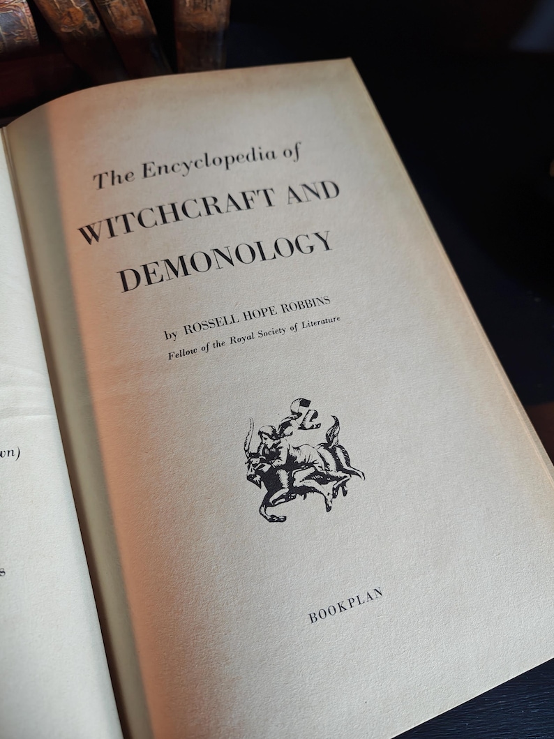 May include: Open book titled "The Encyclopedia of Witchcraft and Demonology" by Rossell Hope Robbins. The book's cover is off-white with black text and a small illustration. The words "BOOKPLAN" are at the bottom.