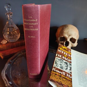 May include: A burgundy-colored book titled "The Encyclopedia of Witchcraft and Demonology" stands upright. A human skull and a crystal decanter are in the background. The book's cover displays the title and author's name.