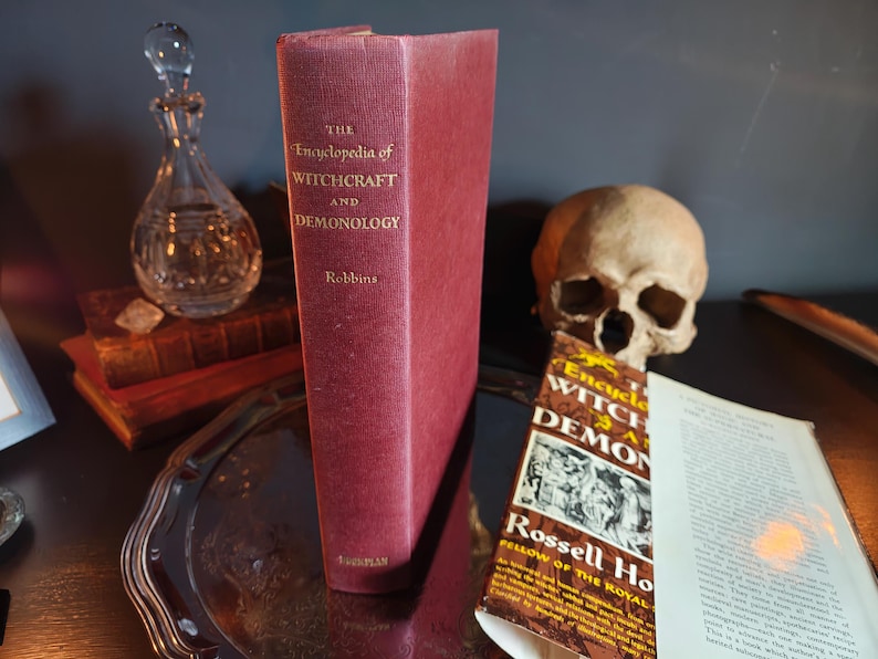 May include: A burgundy-colored book titled "The Encyclopedia of Witchcraft and Demonology" stands upright. A human skull and a crystal decanter are in the background. The book's cover displays the title and author's name.