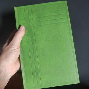 Może przedstawiać: Jasnozielona książka z teksturowaną okładką i wzorem kratki. Książka trzymana jest w dłoni, okładką skierowaną do widza. Krawędzie książki są lekko zużyte, co sugeruje, że jest to książka vintage lub używana.