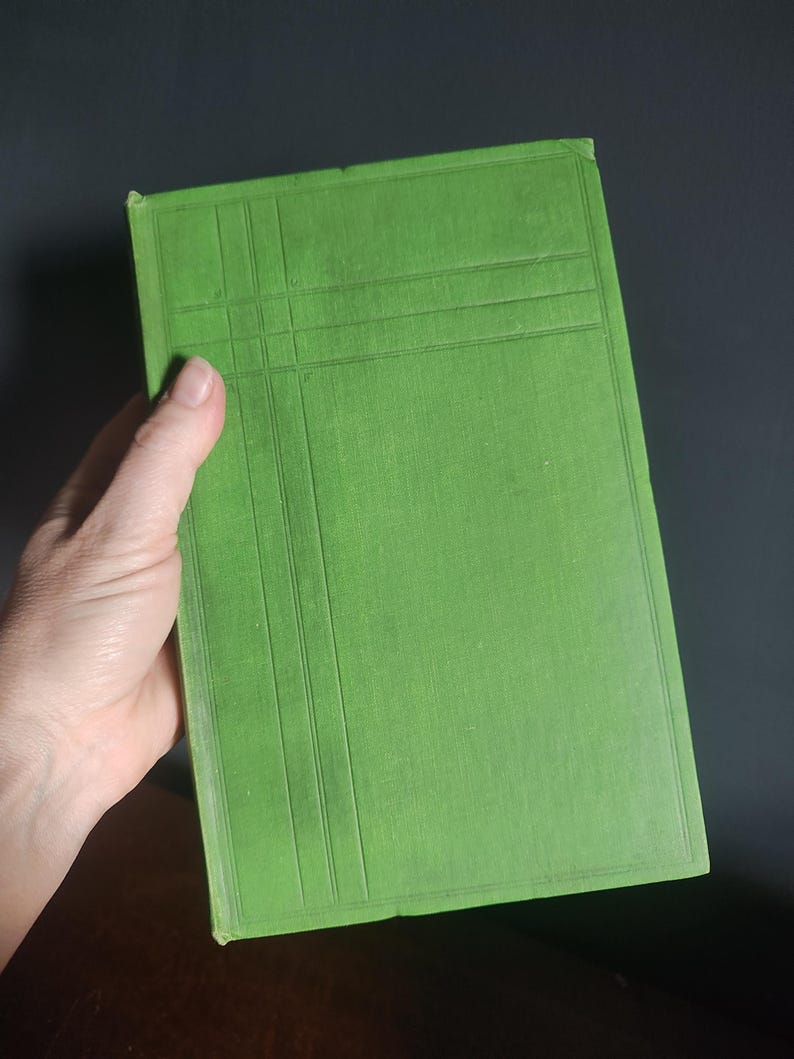 Może przedstawiać: Jasnozielona książka z teksturowaną okładką i wzorem kratki. Książka trzymana jest w dłoni, okładką skierowaną do widza. Krawędzie książki są lekko zużyte, co sugeruje, że jest to książka vintage lub używana.