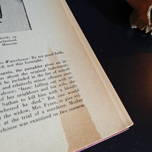 May include: Open book with aged, yellowed pages. Text is visible, with a section of the page stained. The book's spine is red. The text includes the words "Waterhouse" and "indictment."
