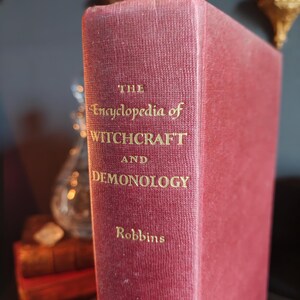 May include: A burgundy-colored book titled "The Encyclopedia of Witchcraft and Demonology" by Robbins. The title and author's name are in gold lettering. The book's spine is visible, with a textured, aged appearance.