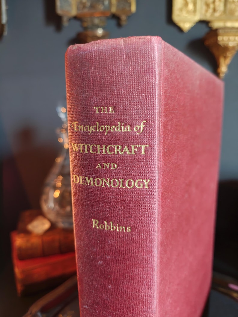 May include: A burgundy-colored book titled "The Encyclopedia of Witchcraft and Demonology" by Robbins. The title and author's name are in gold lettering. The book's spine is visible, with a textured, aged appearance.