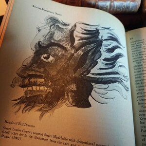 May include: An open book displays a detailed black and white illustration of a demonic face with sharp teeth and wild hair. Text above reads "Aix-en-Provence Nuns" and below "Heads of Evil Demons". The page is aged.