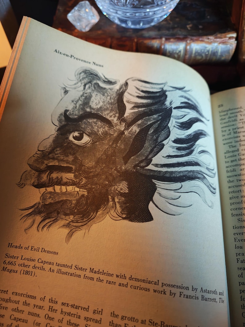 May include: An open book displays a detailed black and white illustration of a demonic face with sharp teeth and wild hair. Text above reads "Aix-en-Provence Nuns" and below "Heads of Evil Demons". The page is aged.