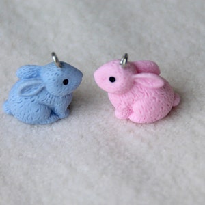 May include: Two small rabbit charms, one light blue and one pink, with silver jump rings. The charms are made of polymer clay and have a fuzzy texture.