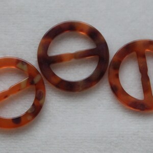 Könnte beinhalten: Drei braune und orangefarbene Gürtelschnallen aus Kunststoff mit einem Mittelbalken. Die Schnallen sind rund und haben ein marmoriertes Muster.