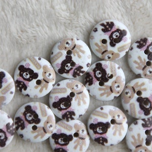 May include: A collection of 15 white wooden buttons with a brown and pink bear design. The bears are holding hands and have a pink bow on their head. The buttons are approximately 1 inch in diameter.