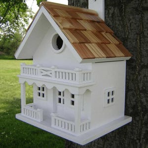 May include: A white wooden birdhouse featuring a natural cedar shingle roof, a circular entrance hole, and a front porch with a balcony railing. The structure includes small square windows and is mounted against a textured tree trunk surface.