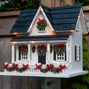 May include: A white wooden birdhouse shaped like a house with a black roof, black shutters, and a white picket fence. The structure is decorated with small green wreaths featuring red bows and is illuminated by a string of warm yellow fairy lights.