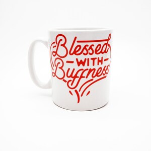 May include: White ceramic mug with a red graphic that says "Blessed with Buffness" in a stylised font.