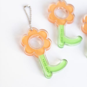 Könnte beinhalten: Ein durchsichtiger orangefarbener Schlüsselanhänger in Form einer Blume mit einem grünen Stiel. Der Stiel trägt den Text "DEAR DAIRA".