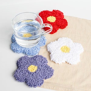 May include: Four crocheted flower-shaped coasters in blue, purple, white, and red with yellow centers. The coasters are on a beige fabric surface with a glass of water on top of the blue coaster.