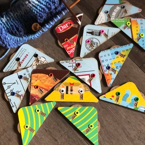 May include: A collection of 11 colorful triangular yarn bowl holders with various designs and text. The bowls are made of cardboard and have a variety of colors, including yellow, green, blue, red, and brown. The text on the bowls includes words like "Kosmos", "Dare", "Humid", and "Shiner".