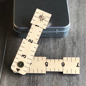 May include: A wooden ruler with markings for inches and a small screw. The ruler is folded into a right angle and has a small yarn ball and knitting needles icon on the top.