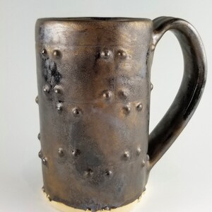 May include: A brown ceramic mug with a textured surface and a gold rim. The mug has a large handle and is decorated with small, raised dots.