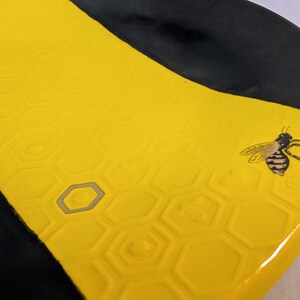 May include: A ceramic dish with a yellow honeycomb pattern and a black border. A detailed illustration of a bee is on the yellow surface. The dish has a glossy finish.