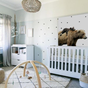May include: A white crib with a white and gray patterned mattress, a wooden baby play gym with hanging toys, and a brown and black bear wall decal with a smaller bear cub on top. The room is decorated with a white rug, a white dresser, and a wooden mobile hanging from the ceiling.