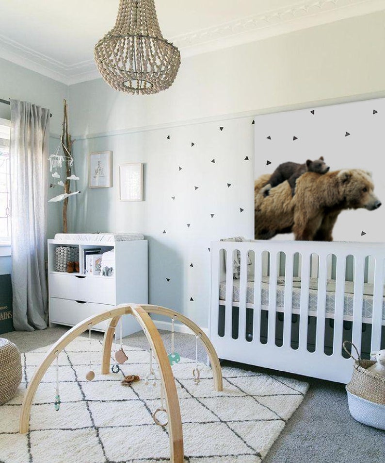 May include: A white crib with a white and gray patterned mattress, a wooden baby play gym with hanging toys, and a brown and black bear wall decal with a smaller bear cub on top. The room is decorated with a white rug, a white dresser, and a wooden mobile hanging from the ceiling.