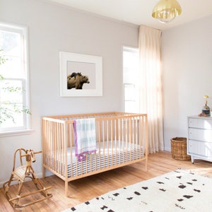 May include: A white crib with a white and blue striped mattress and a light green and purple blanket. A rocking horse is in the foreground. A white dresser with a changing pad is in the background. A woven wall hanging is on the wall behind the dresser. A plant is in a terracotta pot in the corner of the room.