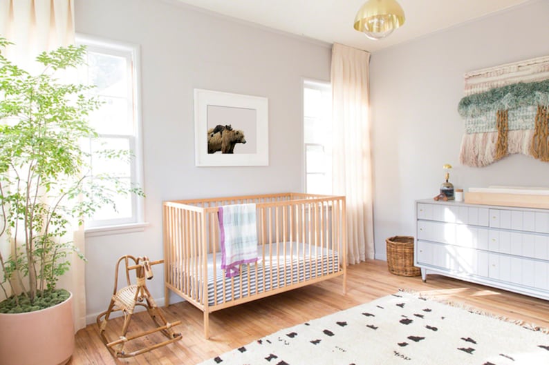 May include: A white crib with a white and blue striped mattress and a light green and purple blanket. A rocking horse is in the foreground. A white dresser with a changing pad is in the background. A woven wall hanging is on the wall behind the dresser. A plant is in a terracotta pot in the corner of the room.