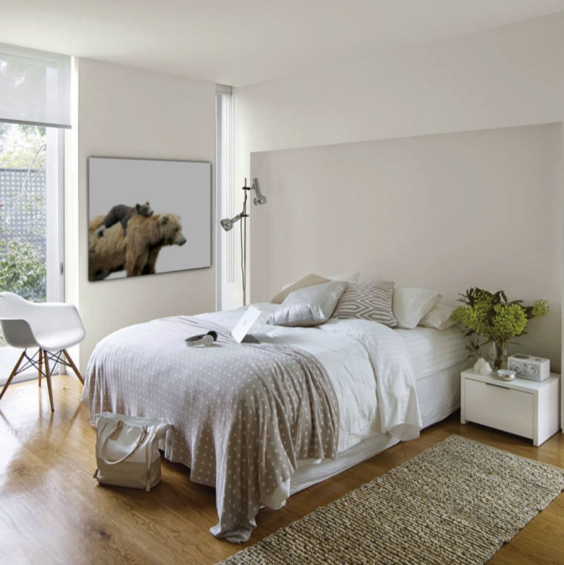 May include: A white bedroom with a bed, a white nightstand, and a framed print of two brown bears. The bed has a white duvet and a gray throw blanket. The nightstand has a small plant and a clock on it.