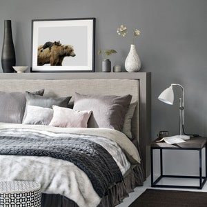 May include: A gray bedroom with a bed, a bedside table, and a framed print of a brown bear and a cub. The bed has a gray headboard and a white and gray blanket. The bedside table has a lamp and a book on it.