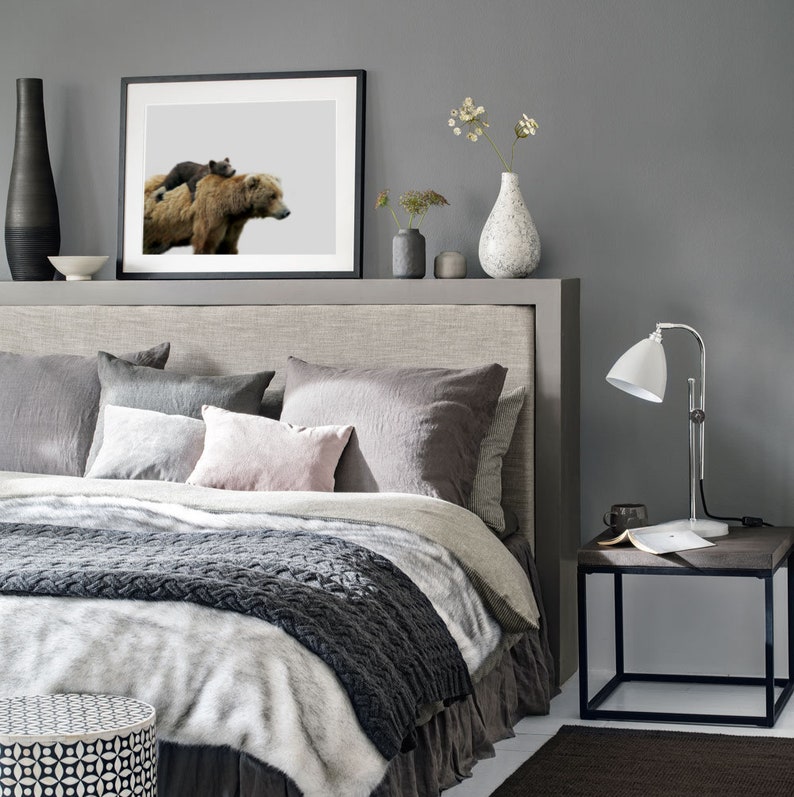 May include: A gray bedroom with a bed, a bedside table, and a framed print of a brown bear and a cub. The bed has a gray headboard and a white and gray blanket. The bedside table has a lamp and a book on it.