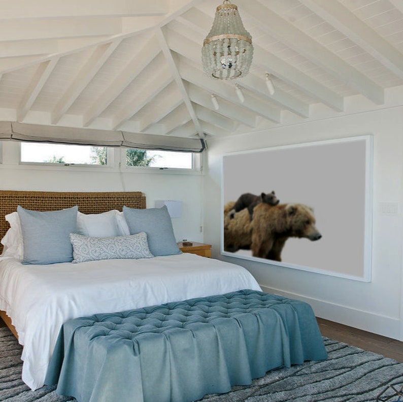 May include: A white bedroom with a large framed print of a brown bear and a cub. The bed has a white duvet and blue pillows. There is a blue ottoman at the foot of the bed.