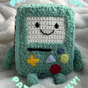 Könnte beinhalten: Ein gehäkeltes Kuscheltier in Form eines Quadrats mit einem lächelnden Gesicht. Das Spielzeug ist hauptsächlich türkis mit einem weißen Gesicht und schwarzen Augen und Mund. Es hat einen gelben Stern, einen roten Kreis, ein blaues Dreieck und einen gelben Kreis auf der Vorderseite. Der Text "PATTERN ONLY!" ist um das Spielzeug geschrieben.