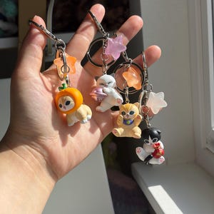 May include: Four metal keychains featuring small cat figurines. One cat wears an orange hat, another holds a pink UFO, a third is orange with a blue collar, and the fourth is black and white holding a red bag. Each includes a star-shaped charm.