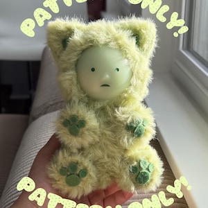 Puede incluir: Una pequeña figura con un rostro liso de color verde pálido y expresión neutra, que lleva una capucha peluda de color verde claro con orejas de gato. El cuerpo está cubierto de pelo largo y suave, con almohadillas de fieltro verde oscuro. Texto: PATTERN ONLY!