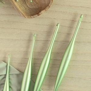 May include: A collection of mint green crochet hooks of varying sizes, arranged on a light wooden surface. The hooks have a smooth, metallic finish and are positioned near a wooden bowl containing additional hooks. The image is well-lit, highlighting the tools.