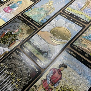 May include: A collection of tarot cards with gold foil accents and intricate illustrations. The cards feature images of figures, symbols, and numbers, with a focus on the "One of Pentacles" card. The cards are arranged on a textured surface.