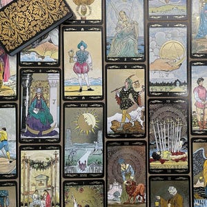 May include: A collection of tarot cards with gold and black borders, featuring various illustrations and symbols. The cards are arranged on a surface, with a decorative box in the upper left corner. The cards depict figures, celestial bodies, and other symbolic imagery.