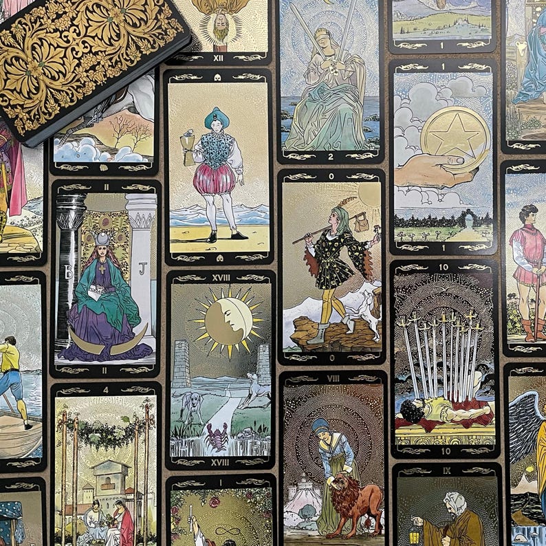 May include: A collection of tarot cards with gold and black borders, featuring various illustrations and symbols. The cards are arranged on a surface, with a decorative box in the upper left corner. The cards depict figures, celestial bodies, and other symbolic imagery.